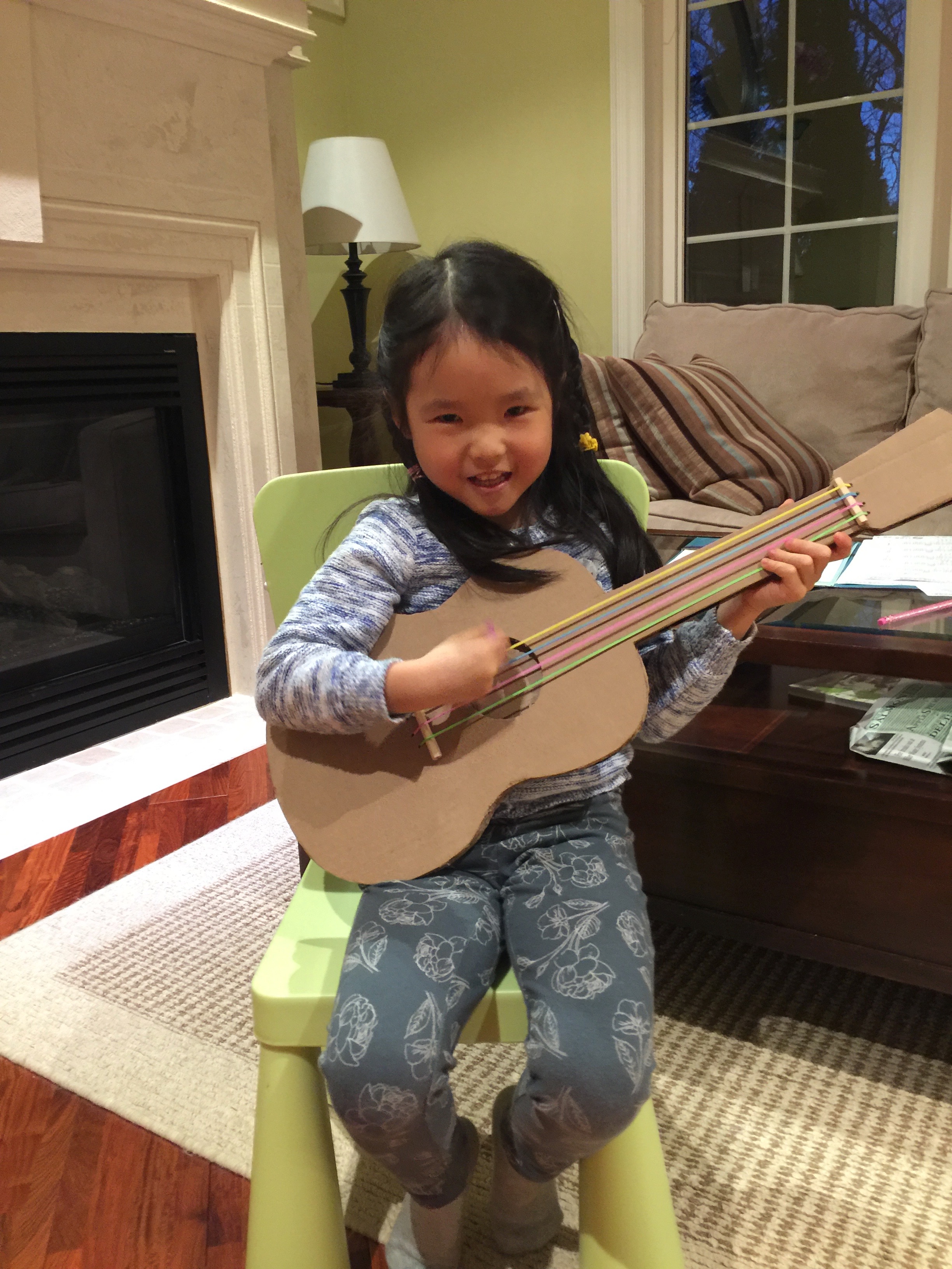 A young Mia smiling and playing a handmade cardboard guitar craft with rubber-band strings