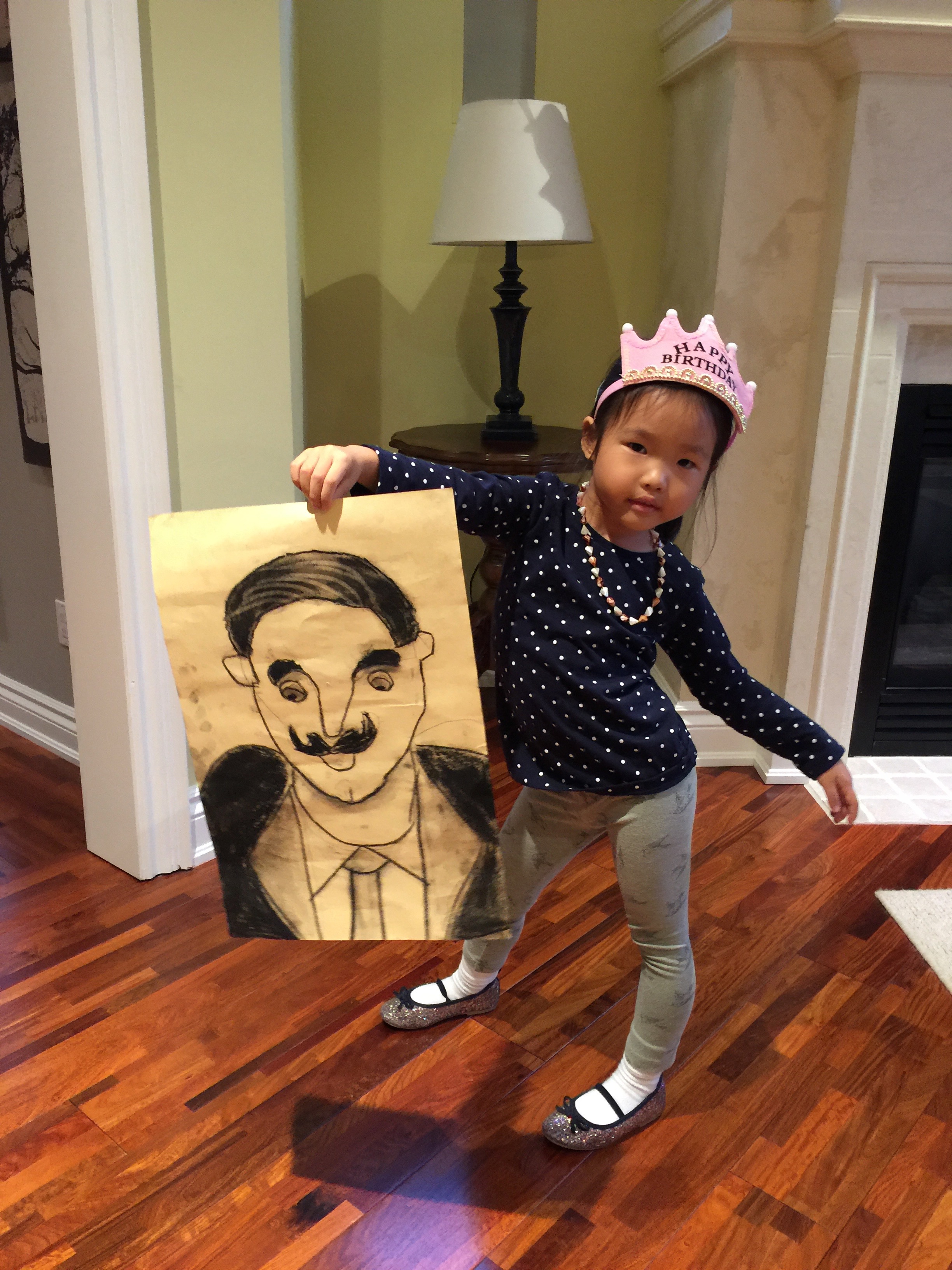 A young Mia in a birthday crown holding up a charcoal portrait drawing of a man with a mustache