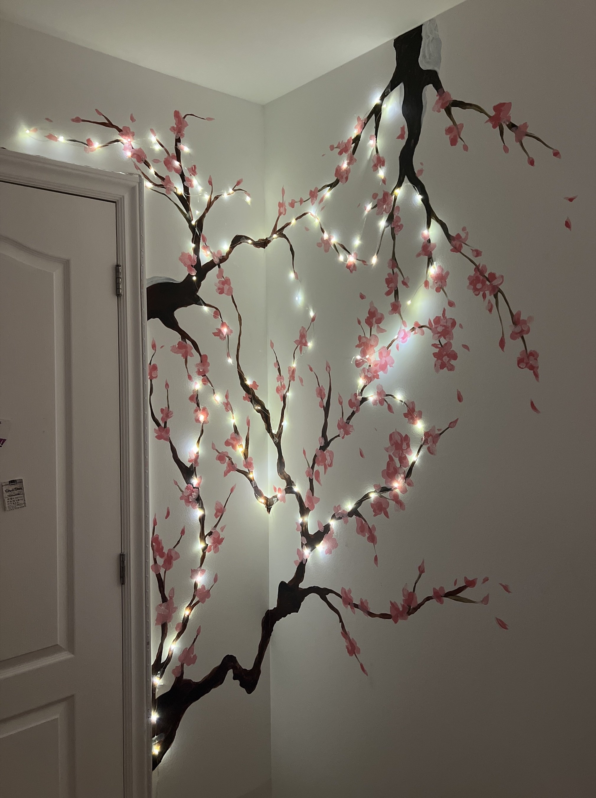 A hand-painted cherry blossom branch mural climbing a wall corner, woven through with string lights