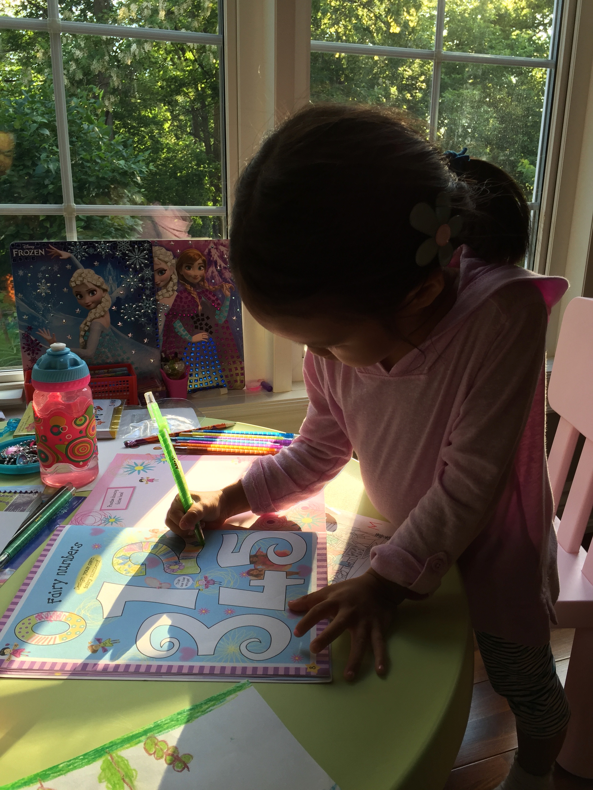 A young Mia coloring in a fairy-themed numbers activity book by a window