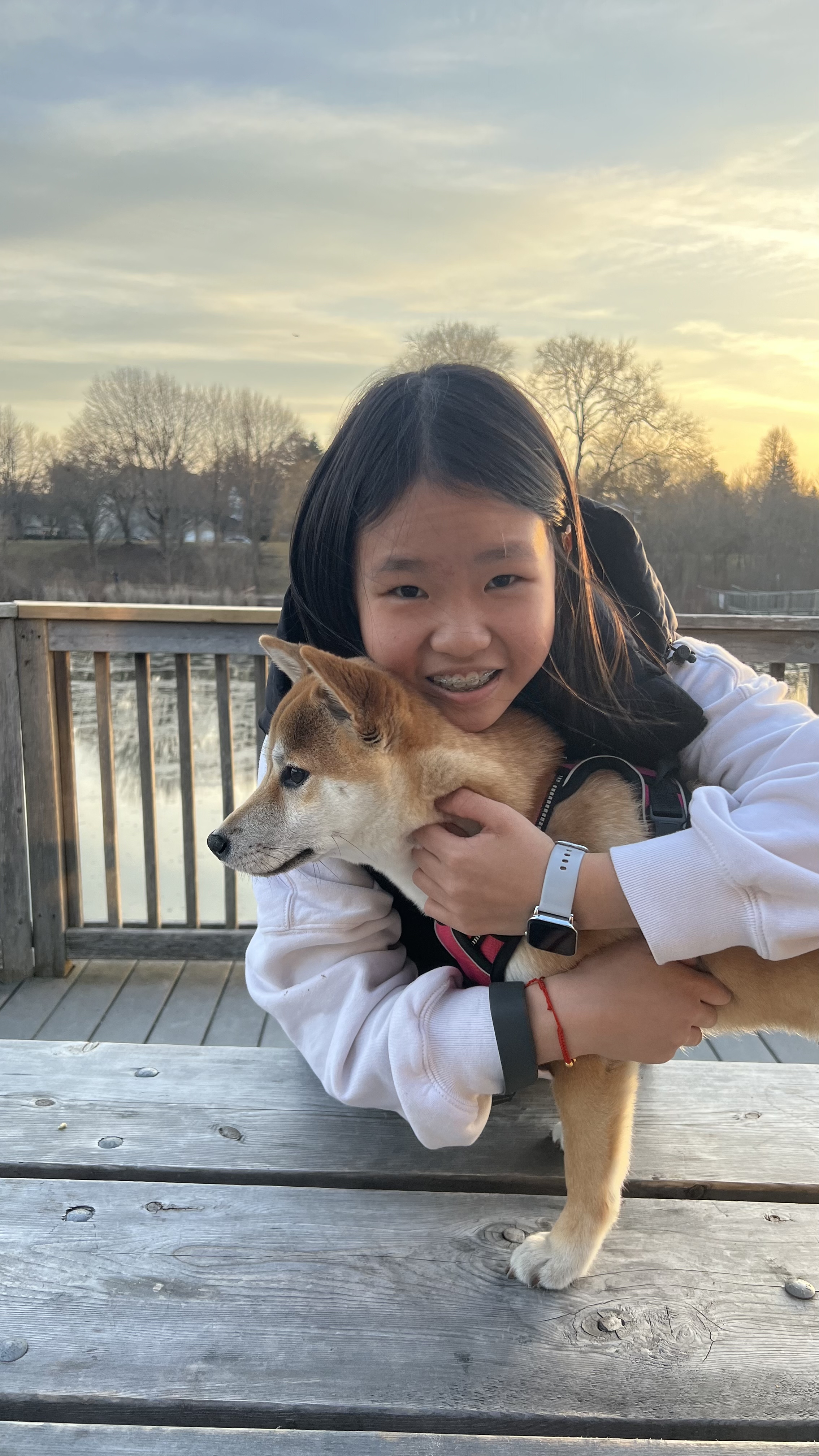 Mia holding her Shiba Inu on a dock at sunset, with a pond behind them