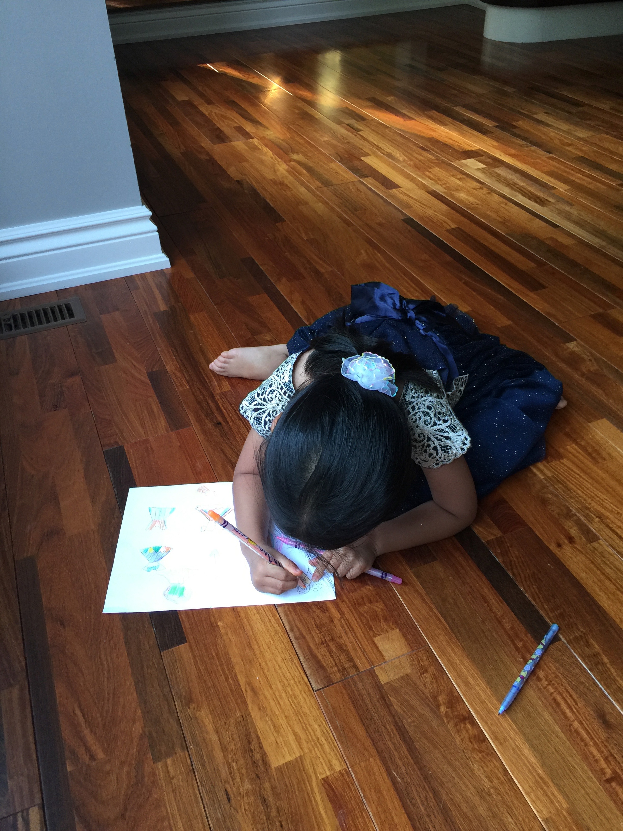 A young Mia in a party dress lying on the floor coloring a drawing