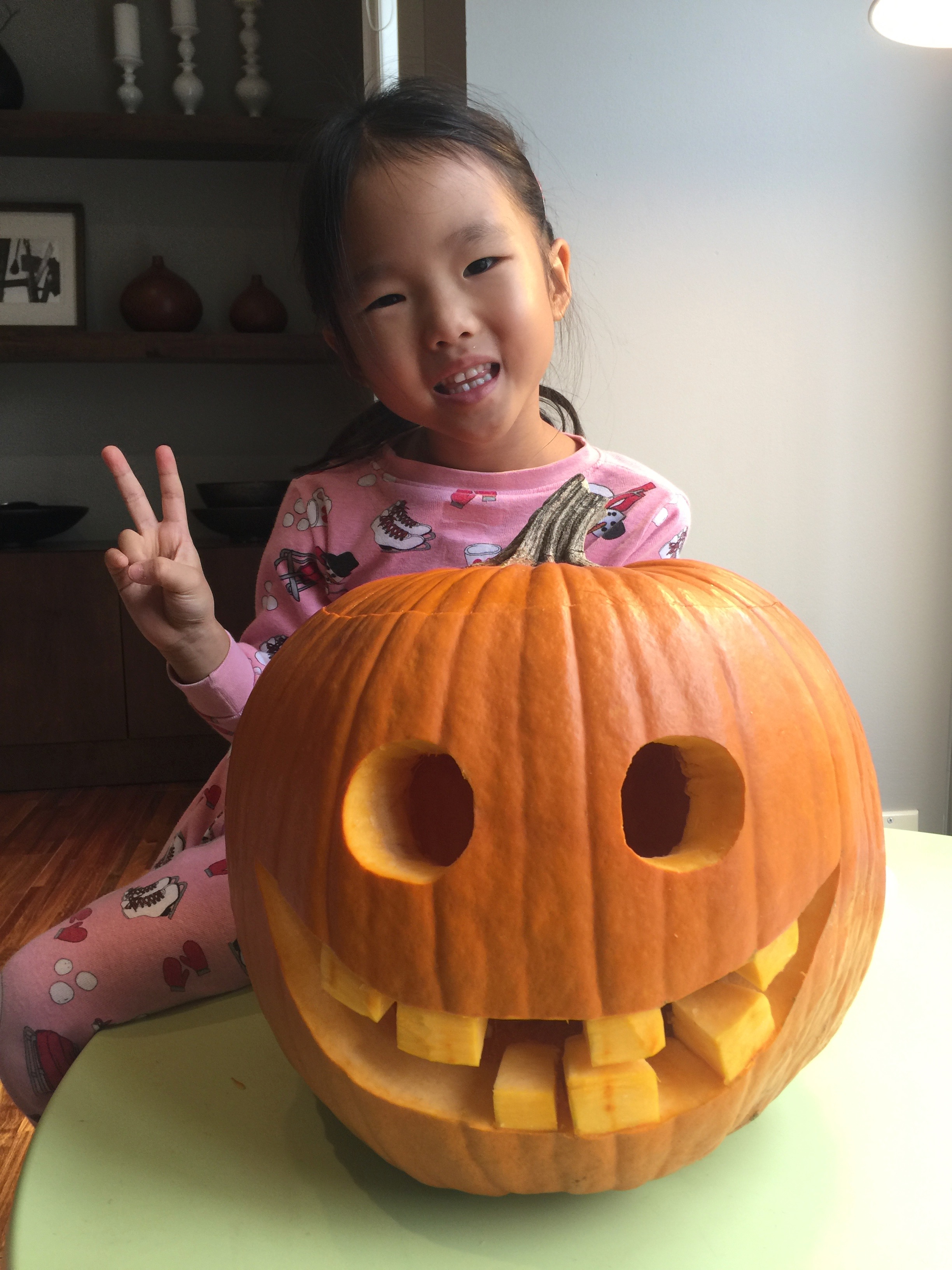 A young Mia making a peace sign next to a carved jack-o'-lantern at home