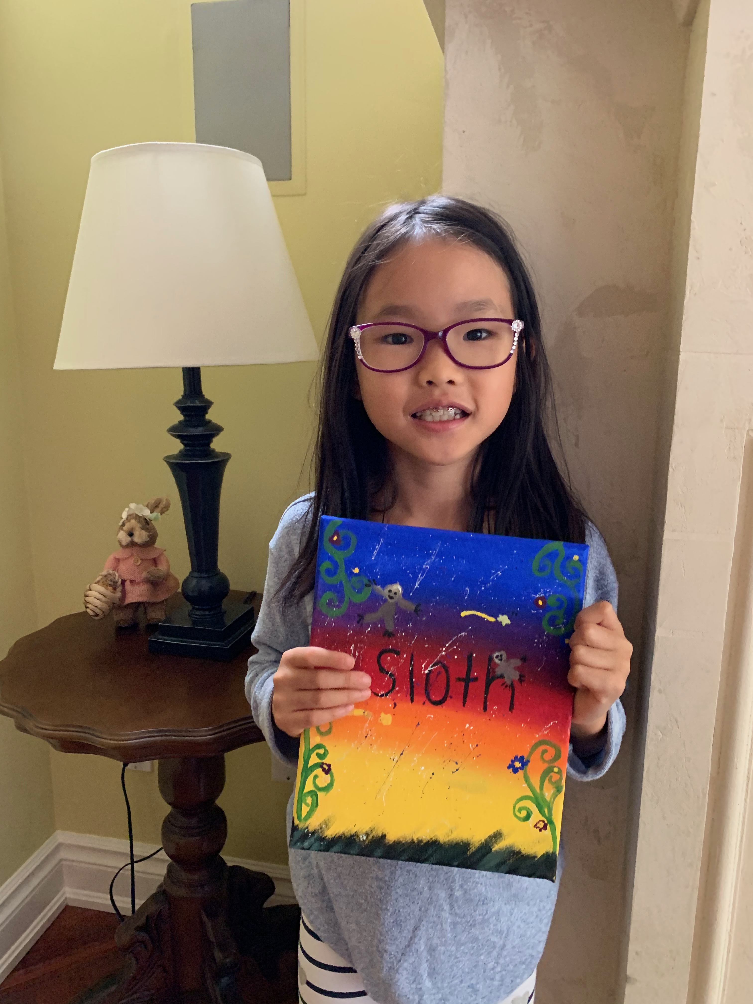 A young Mia smiling and holding her sunset-toned painting of a sloth titled "Sloth"
