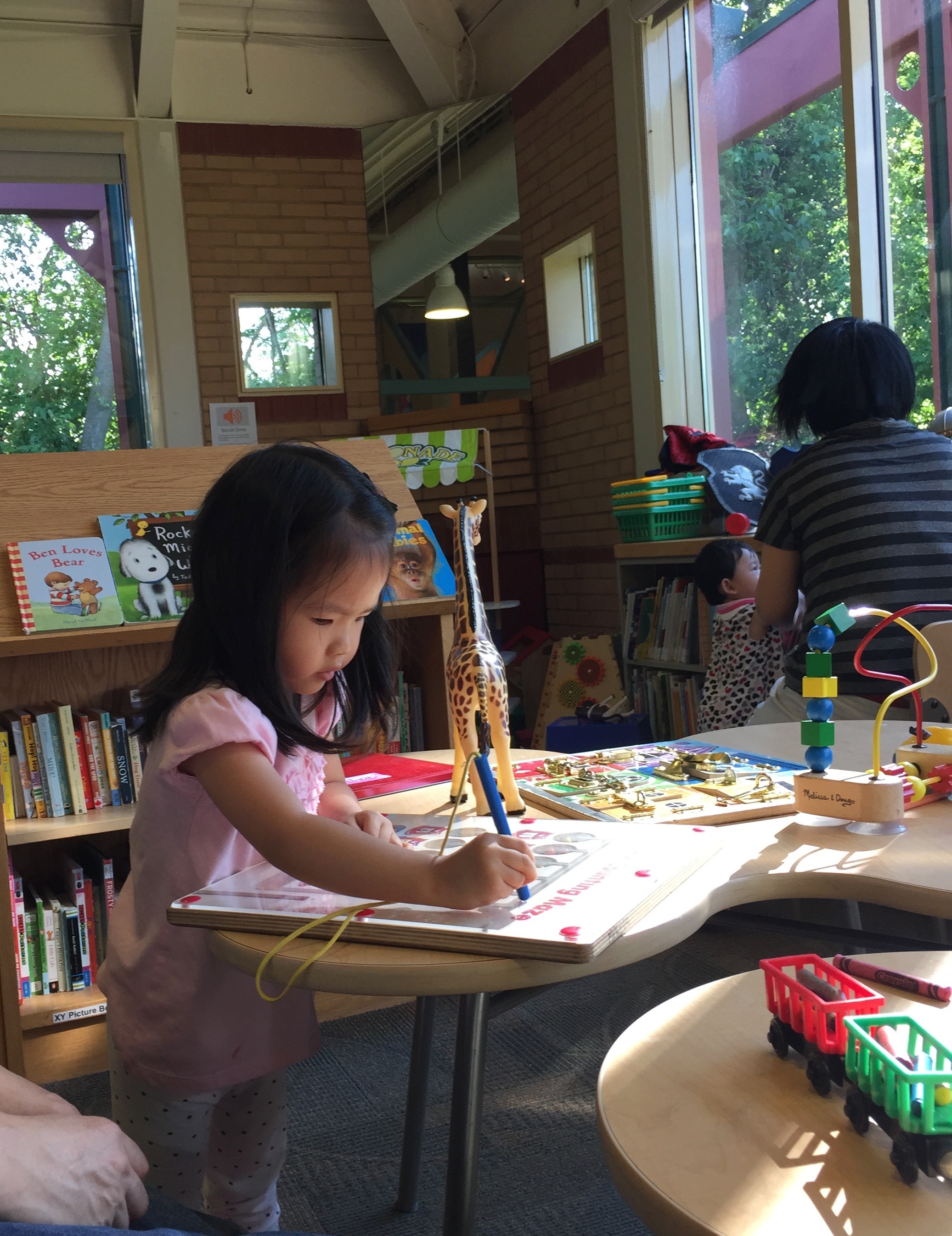 A young Mia at a children's activity table in a library, drawing on a wooden maze board next to a toy giraffe