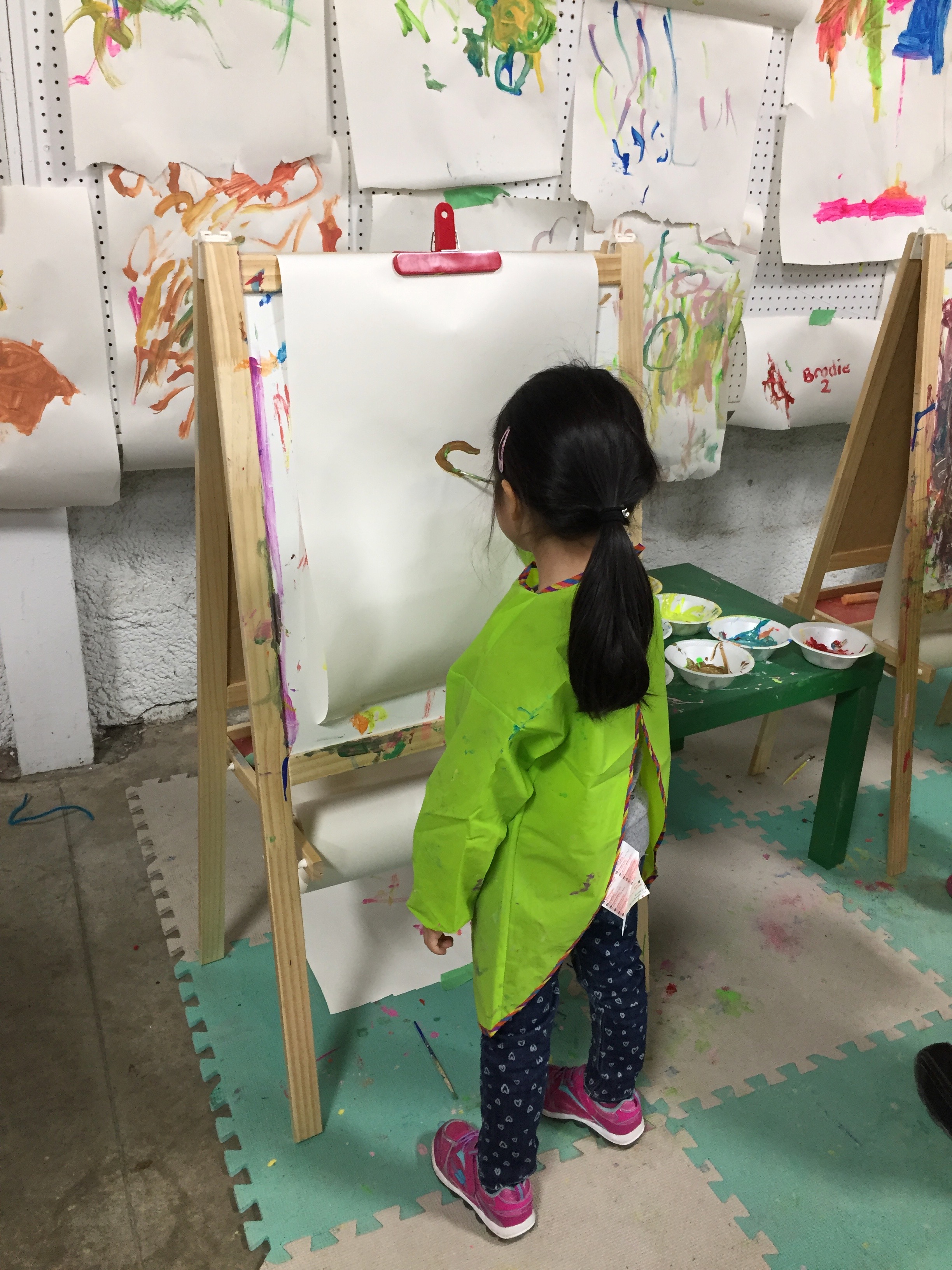 A young Mia in a painting smock at an easel in an art studio, surrounded by other children's paintings on the walls