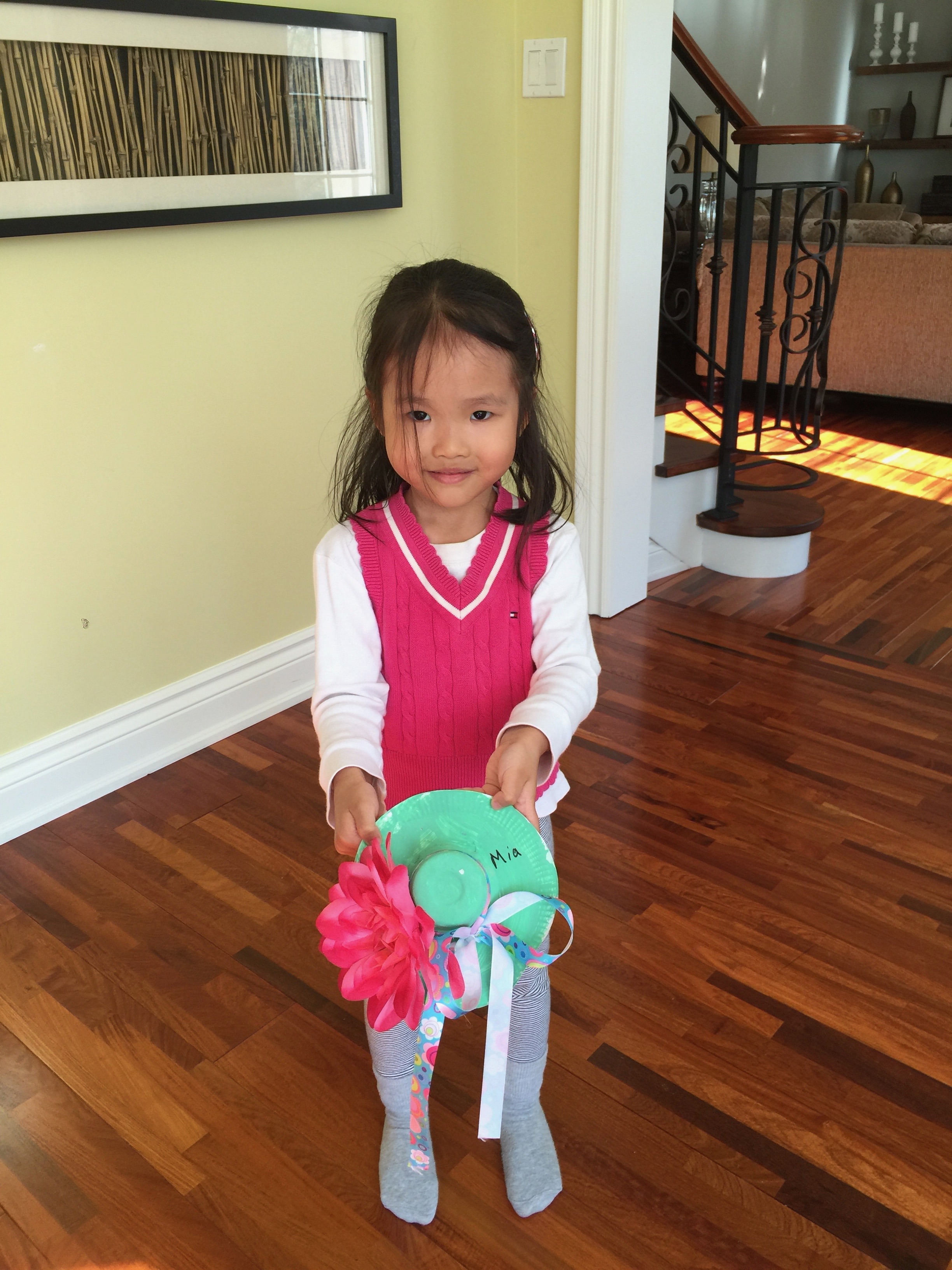 A young Mia holding a paper-plate craft hat decorated with a flower and ribbon, with her name written on it