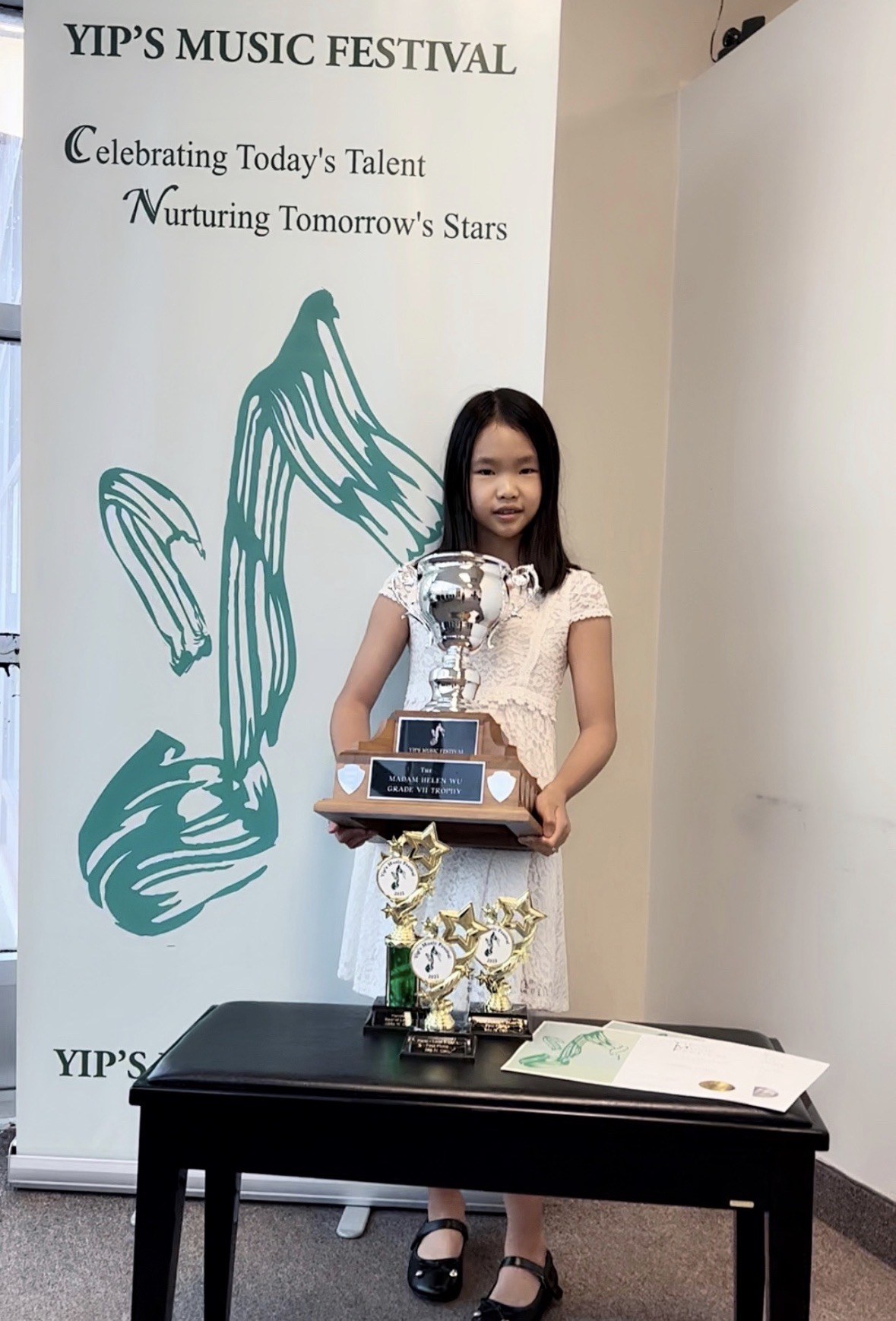 Mia holding the Madam Helen Wu Grade VII Trophy at Yip's Music Festival, with two more piano trophies on the table beside her