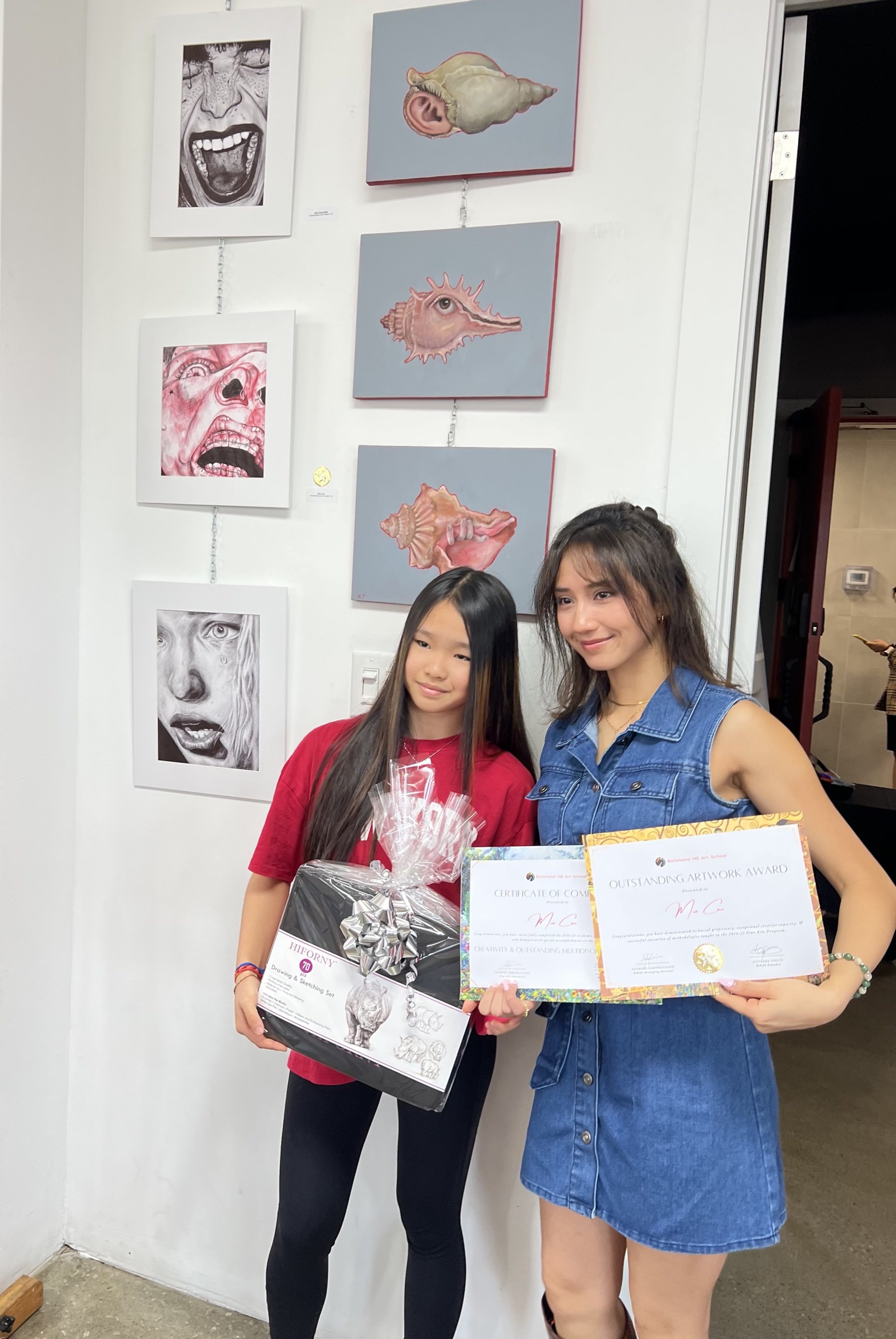 Mia holding an Outstanding Artwork Award and certificate of completion with her art teacher, in front of a wall of her framed drawings and paintings