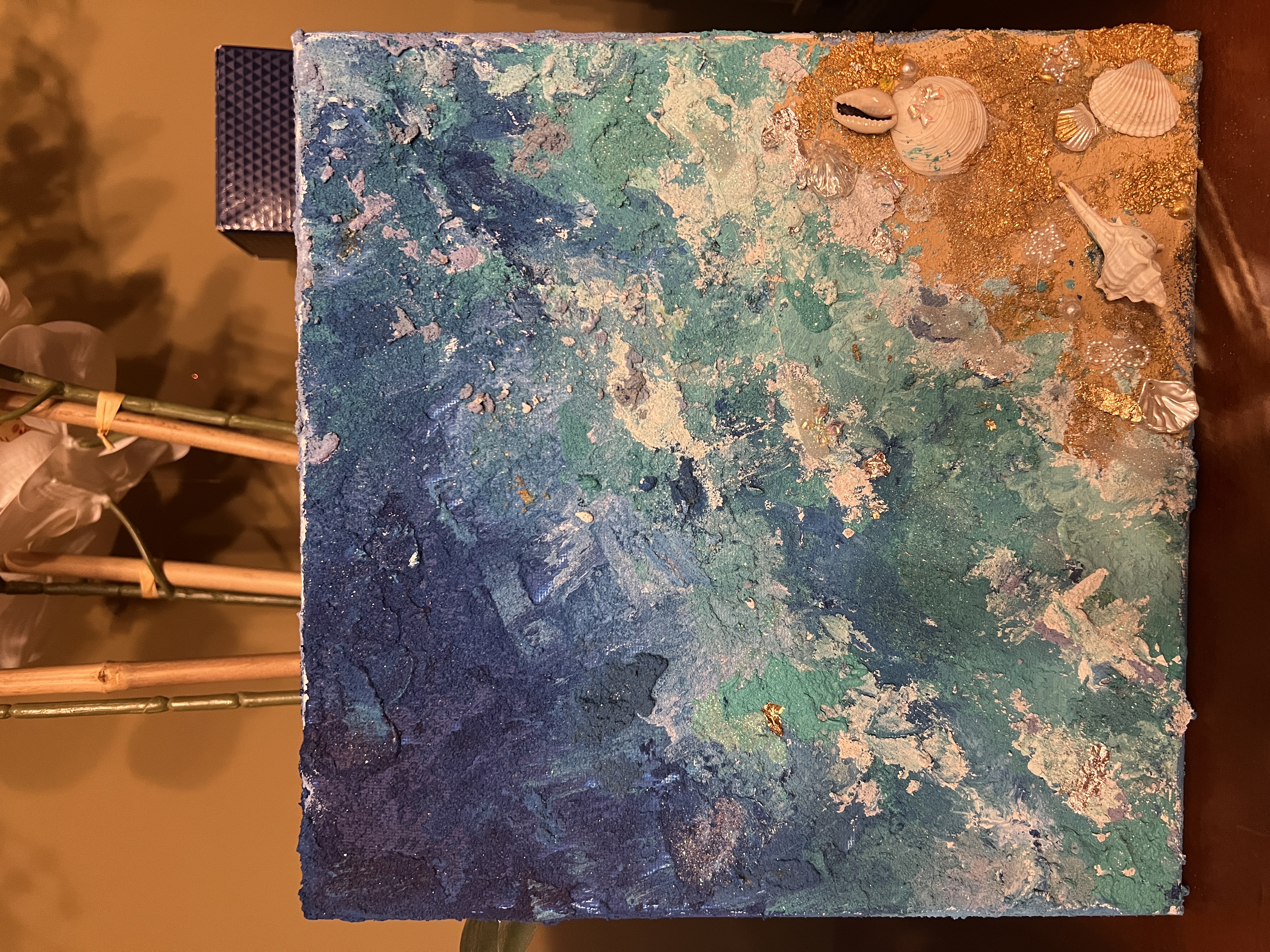 A textured mixed-media abstract ocean painting in blue and green with real seashells embedded in gold sand