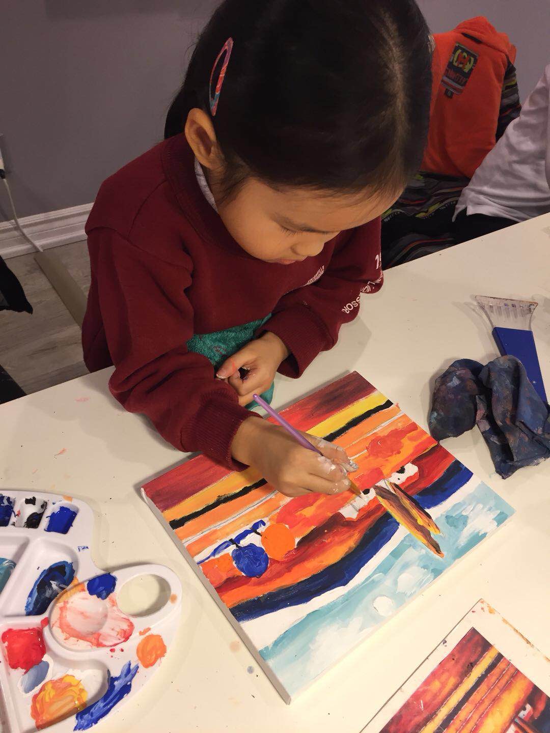 A close-up of young Mia painting an abstract piece in orange, black, and blue stripes