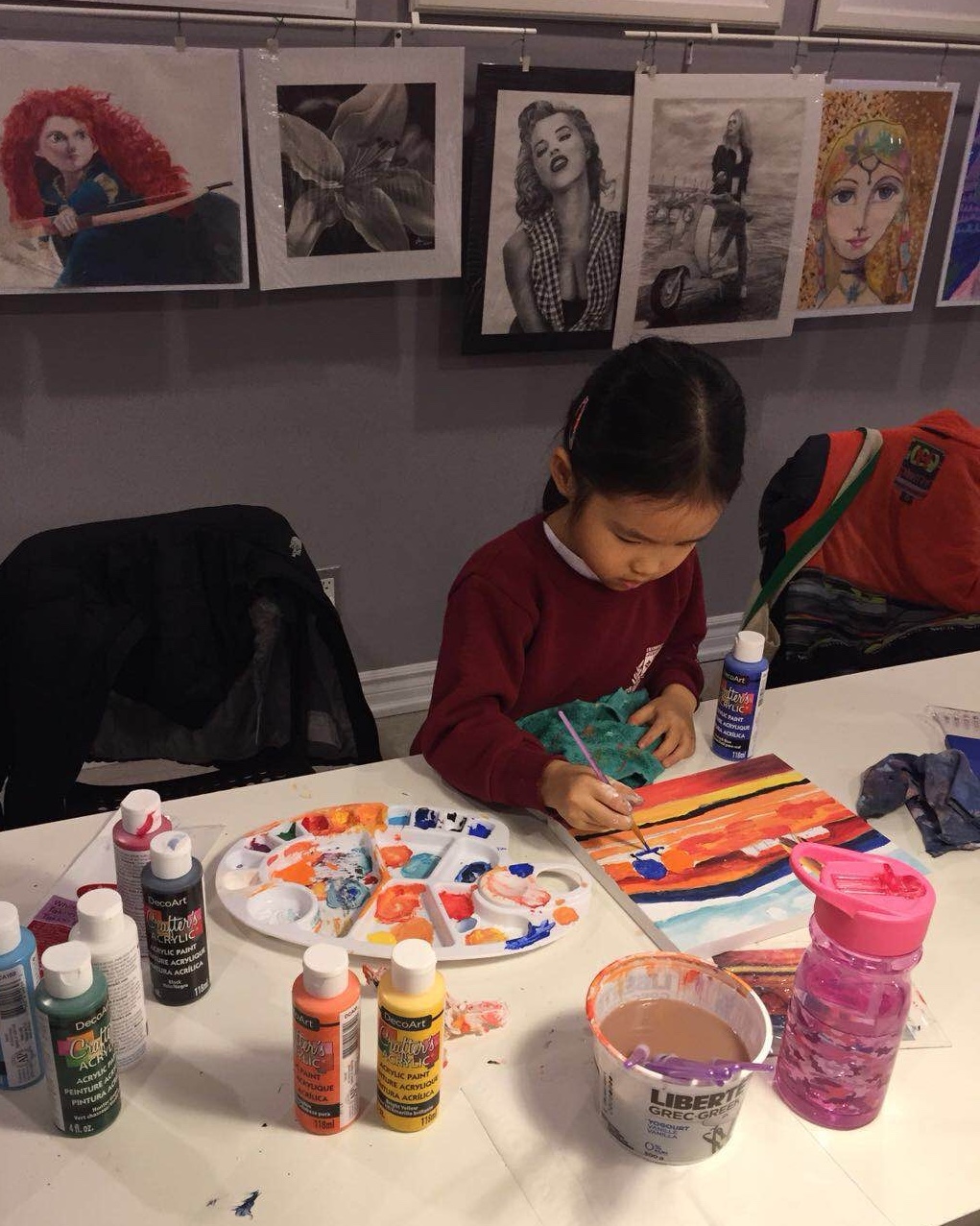 A young Mia painting a sunset scene at a table in an art class, with framed portrait paintings hanging on the wall behind her