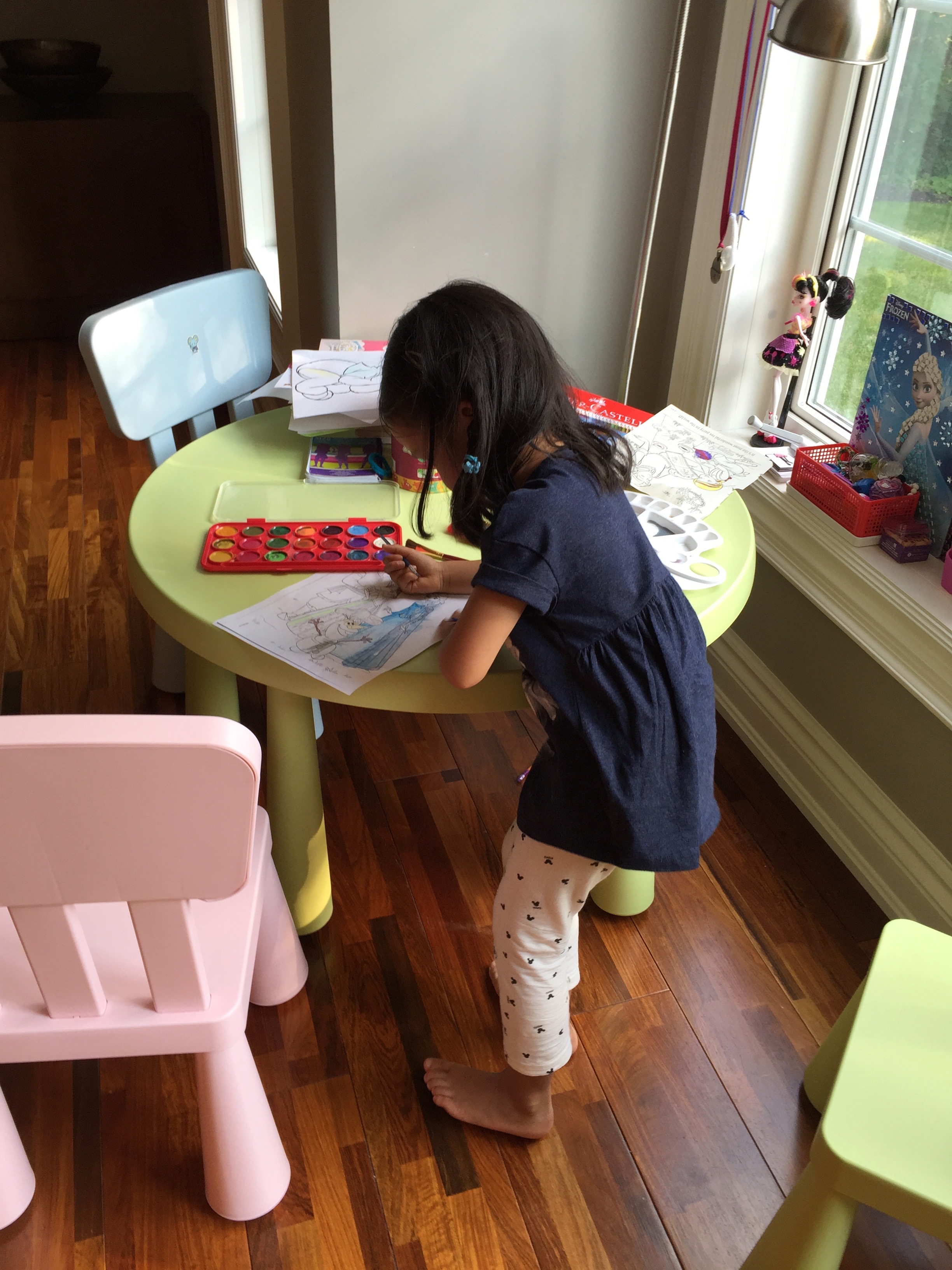 A young Mia painting with a watercolor palette at a small table by a window